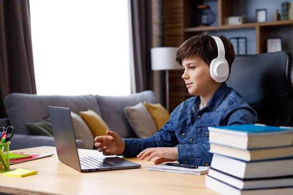 Schoolboy wearing earphones has online lesson, video call with teacher, answering questions, check of knowledge. Cute teenager studying at home using laptop. Distance learning and home education.
