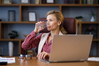Ofiste dizüstü bilgisayarla çalışan bitkin bir iş kadını. Stresli bir kadın bir bardak su içip içiyor. Sağlık hizmeti ve refah yaşam tarzı. İçme suyu. Diyet konsepti. Sağlıklı beslenme. Susuzluk..