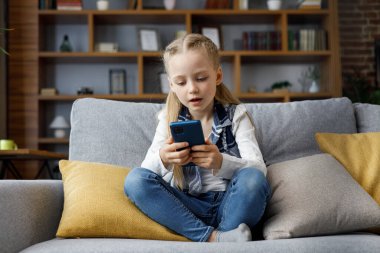 Akıllı telefon tutan ve evdeki kanepede dinlenen şirin küçük kız portresi. Favori müzik veya karikatür seçimi, mesajlaşma, internette gezinme, video izleme, cep telefonunda oyun oynama.