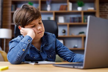 Young boy studying remotely at home using laptop. Tired schoolboy falling asleep during an online lesson with teacher. Distance learning is boring and uninteresting.