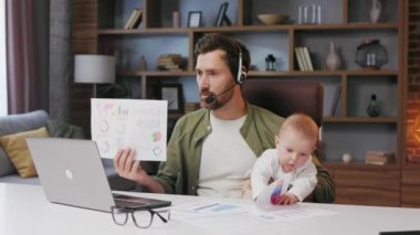 Father on maternity leave, holding an online conference, a meeting in the home office with a small child in his arms. Dad with a child. Freelancer combines child care and work. Paternity leave.