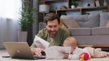 Dad is on maternity leave, he works lying on a mat in an apartment with a laptop and a child. Father care for baby. Father work with child at home. Paternity Leave