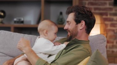 Dad plays with his little baby, on the couch in a cozy apartment. Fatherly love. Father hugs and kisses his little daughter.