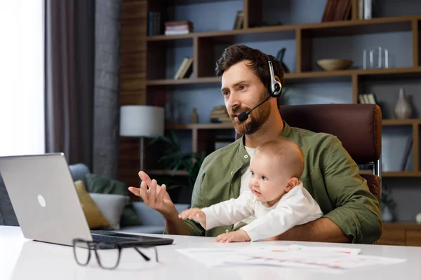 Father on maternity leave, holding an online conference, a meeting in the home office with a small child in his arms. Dad with a child. Freelancer combines child care and work. Paternity leave.