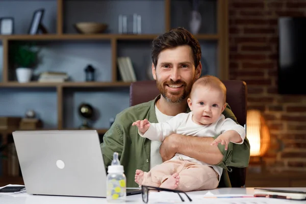 Portrait of a Father on maternity leave, in his home office with a small child in his arms. Dad at the table with a laptop and a child. Holiday to care for the child. Maternity leave for a man