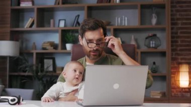 Father is on maternity leave, working in a home office at a laptop with a small child in his arms. Freelance businessman combines child care and work. Paternity Leave