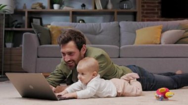 Dad is on maternity leave, he works lying on a mat in an apartment with a laptop and a child. Father care for baby. Father work with child at home. Paternity Leave