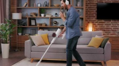 Cheerful guy, the man cleans the apartment. Vacuuming, wearing headphones with dancing and singing into a smartphone instead of a microphone. House cleaning is easy