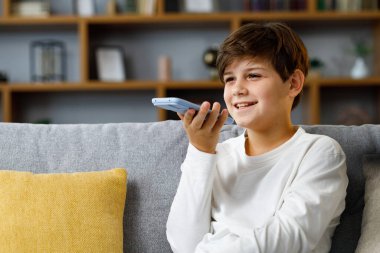 Young cute boy recording voice message on smartphone. Smiling teenager using virtual assistant app sitting at coach at home. Voice recognition, audio messages, using translator on mobile phone.