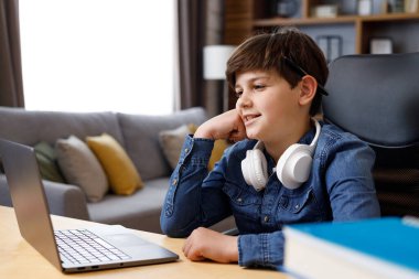 Portrait of cute schoolboy looking at laptop screen and smiling. Teenager studying remotely at home using laptop. Distance learning and home education. Happy childhood.
