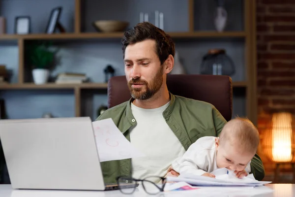 Father on maternity leave, holding an online conference, a meeting in the home office with a small child in his arms. Dad with a child. Freelancer combines child care and work. Paternity leave.