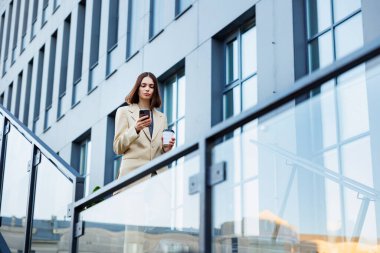 Genç bir esmer kız bir iş merkezinin arka planına karşı, ofis merkezine. Başarılı bir girişimin portresi. Akıllı telefon, kahve kıyafeti.