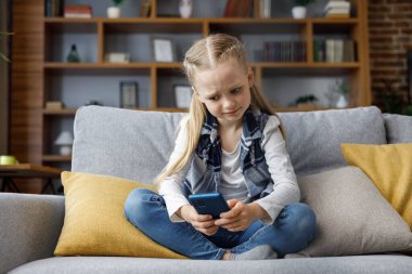 Portrait of cute little cute girl holding smartphone and resting at sofa at home. Choosing favorite music or cartoons,texting messages,browsing internet,watching video, playing games on mobile phone.