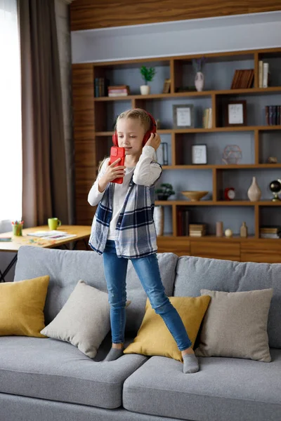 Happy little girl wearing red earphones dancing and singing using smartphone like microphone. Cute child having fun jumping on coach. Home leisure, happy kid. Attention deficit hyperactivity disorder