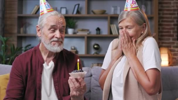 一对头戴圆锥帽的白发苍苍的老夫妇在家里欢度生日 有爱心的丈夫拥抱妻子 庆祝结婚周年 吹灭生日蛋糕上的蜡烛 — 图库视频影像