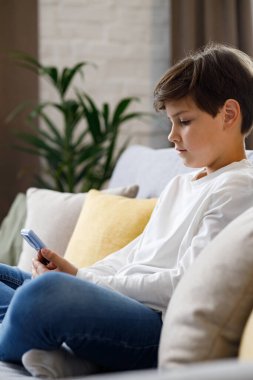 Portrait of young cute boy holding smartphone sitting at sofa. Teenager at home choosing favorite music, texting messages, browsing internet, watching video in social media, playing online games