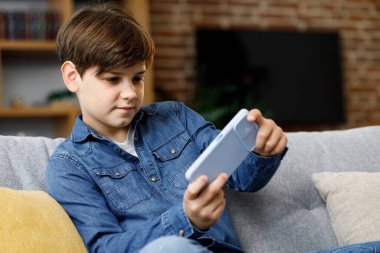 Cheerful young boy playing video game on smartphone sitting on sofa at home. Cute teenager playing in car racing game on mobile phone. Spending time at home, leisure, relax, happy carefree childhood.