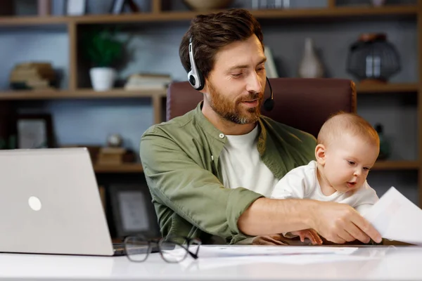 Father on maternity leave, holding an online conference, a meeting in the home office with a small child in his arms. Dad with a child. Freelancer combines child care and work. Paternity leave.