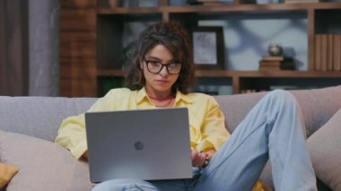 Evden bağımsız olarak çalışan ciddi, genç bir kadın. Kanepede dizüstü bilgisayarda çalışıyor, bilgisayarla evde çalışıyor, kadın kullanıcı, internet uzaktan kumandası, programlama.