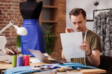 A young guy is a fashion designer. He looks at the sketch while sitting at a table with a sewing machine.