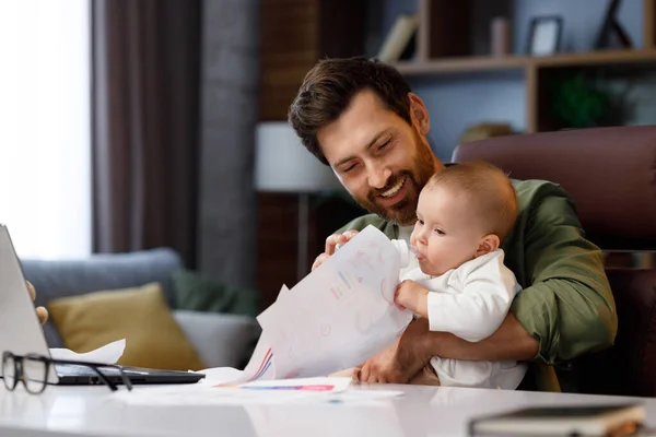 Father is on maternity leave, working in a home office at a laptop with a small child in his arms. Freelance businessman combines child care and work. Paternity Leave