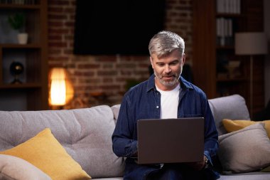 Dizüstü bilgisayar kullanan serbest çalışan bir erkek uzaktan çalışıyor ve kanepede oturuyor. Evde uzaktan çalışırken dizüstü bilgisayardaki video konferansına katılan odaklanmış bir adam.