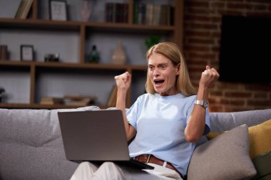 Dizüstü bilgisayara bakıp başarıyı kutlayan, evdeki koltukta oturan heyecanlı kadın. Mutlu bayan, elinde bilgisayarla harika haberler alıyor, yeni bir iş fırsatı ve motive olmuş hissediyor..