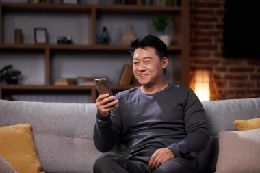 Gülümseyen Asyalı adam sosyal ağlarda iletişim kuruyor ve akıllı telefon kullanıyor. Sıradan giyinmiş bir adam cep telefonundan bakar, dostça iletişim kurar, kanepeye oturur ve evde dinlenir.