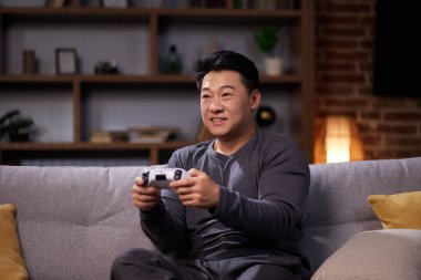 Kablosuz kumanda kullanan ve evde video oyunu oynayan mutlu bir Asyalı. PlayStation kullanan ve kanepede dinlenirken sanal oyunlarda yarışan neşeli bir adam..