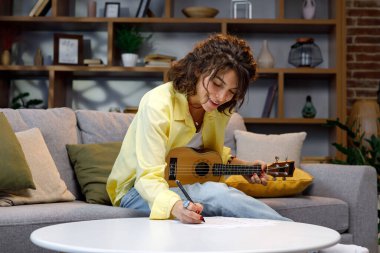 Sarı tişörtlü güzel bir hippi kız evdeki kanepede gitar çalıp şarkı söylüyor. Bir şarkı besteler, notlar yazar. Evde müzik aletleri çalan hobiler