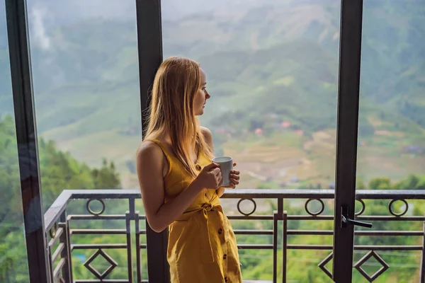 Una giovane donna che tiene in mano una tazza di caffè mentre siede sulla sedia sul balcone, guardando le montagne e la natura verde. Donna a Sapa nella nebbia, Vietnam nord-occidentale. Vietnam concetto di viaggio. Patrimonio UNESCO — Foto Stock