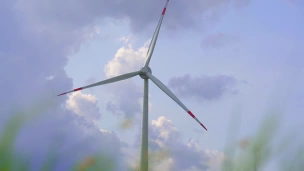 Colpo in mano. Una turbina eolica con un cielo blu nuvoloso sullo sfondo. Vista attraverso l'erba. Investimenti in energia verde. Concetto di elettricità verde — Video Stock