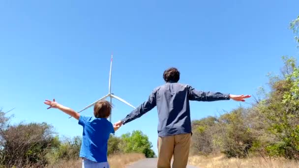Fotografía en cámara lenta de un padre y un hijo corriendo por una carretera que conduce al generador eléctrico eólico. Inversión en energía verde. Concepto de electricidad verde — Vídeos de Stock