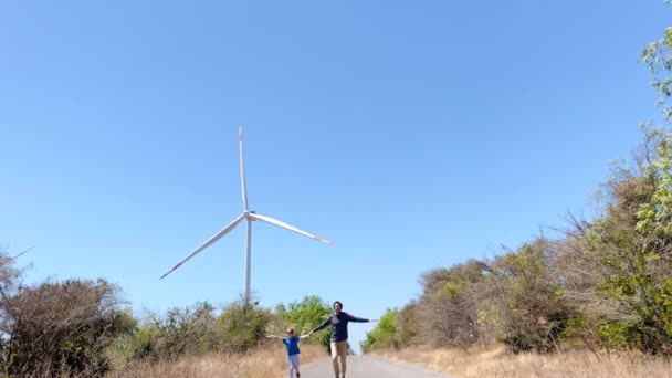 Filmagem lenta de um pai e filho correndo em uma estrada que leva ao gerador de energia eólica. Investimento em energia verde. Conceito de electricidade verde — Vídeo de Stock