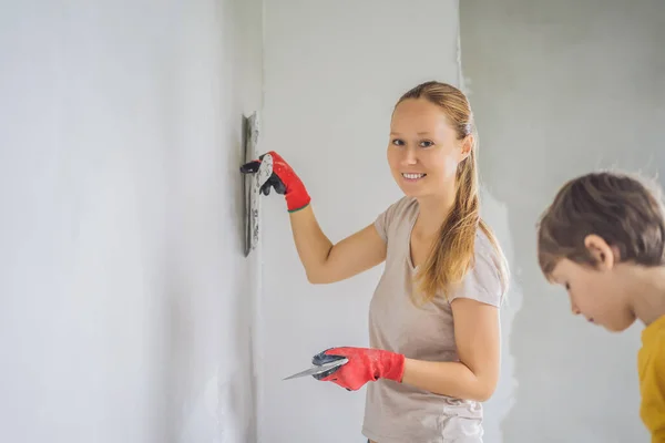 Donna con suo figlio fa riparazioni a casa, insegna al ragazzo a intonacare le pareti con una spatola in mano. — Foto Stock