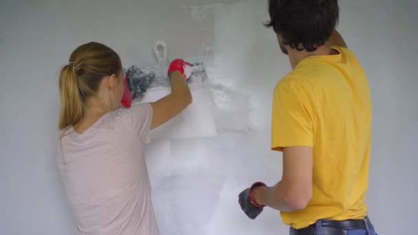 Молодий чоловік і жінка роблять ремонт стін у своєму будинку. Вони наносять шпаклівку на стіну. Зробити ремонт самостійно — стокове відео