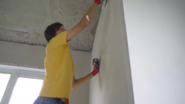 黄色のTシャツを着た若い男が家の壁の修復をしている。彼は壁にパテを塗っている。自分で改装しろ — ストック動画