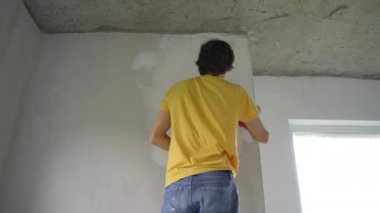Sarı tişörtlü genç bir adam evinde duvar tadilatı yapıyor. O, macunu duvara uyguluyor. Tadilatı kendin yap.