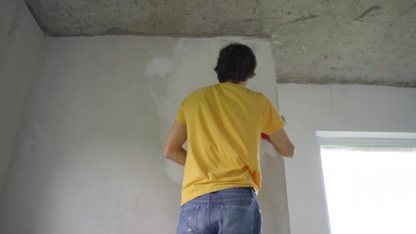 Un joven con una camiseta amarilla está haciendo una renovación de paredes en su casa. Está aplicando masilla a la pared. Haga la renovación usted mismo — Vídeo de stock