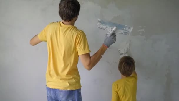 Un joven y su hijo están haciendo una renovación de paredes en su casa. Están aplicando masilla a la pared. Haz la renovación tú mismo. Enseñar a los niños cómo hacer las reparaciones por su cuenta. Ellos están teniendo una — Vídeos de Stock