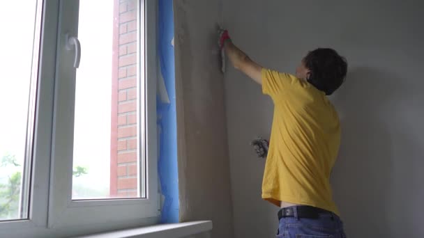 Молодой человек в жёлтой футболке делает ремонт стен в своём доме. Он накладывает шпатлевку на стену. Сделайте ремонт самостоятельно — стоковое видео