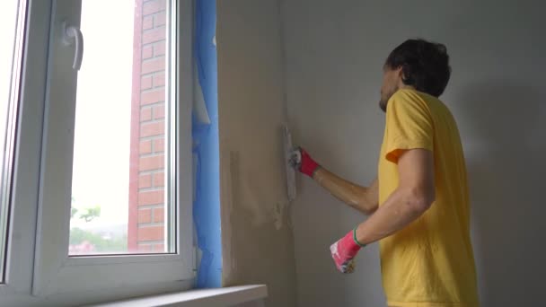 Молодий чоловік у жовтій футболці робить ремонт стін у своєму будинку. Він наносить шпаклівку на стіну. Зробити ремонт самостійно — стокове відео