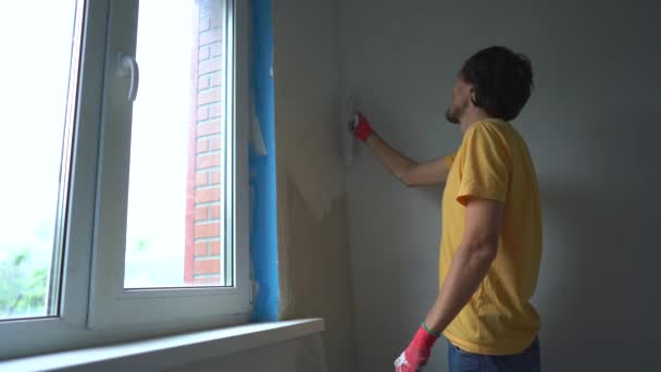 Молодий чоловік у жовтій футболці робить ремонт стін у своєму будинку. Він наносить шпаклівку на стіну. Зробити ремонт самостійно — стокове відео