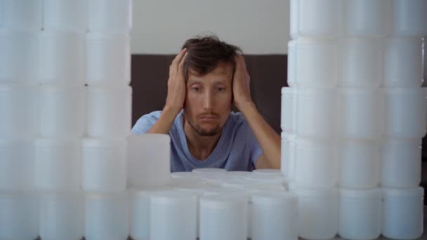 Un hombre está sentado frente a una pared hecha de latas de plástico. Está horrorizado por la cantidad de plástico que tiene que disponer. Detener la contaminación plástica. Reducir los plásticos de un solo uso — Vídeos de Stock