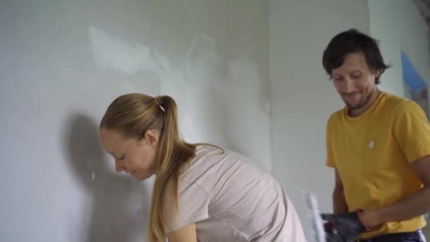 Молодий чоловік і жінка роблять ремонт стін у своєму будинку. Вони наносять шпаклівку на стіну. Зробити ремонт самостійно — стокове відео