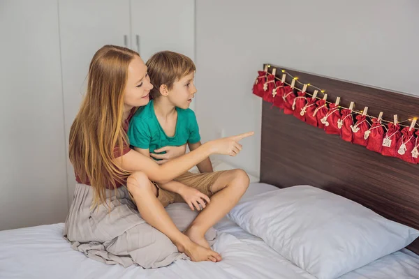 Muchacho sonriente sentado en una cama abriendo el primer regalo del calendario de adviento. Tradición estacional de invierno. Copiar espacio — Foto de Stock