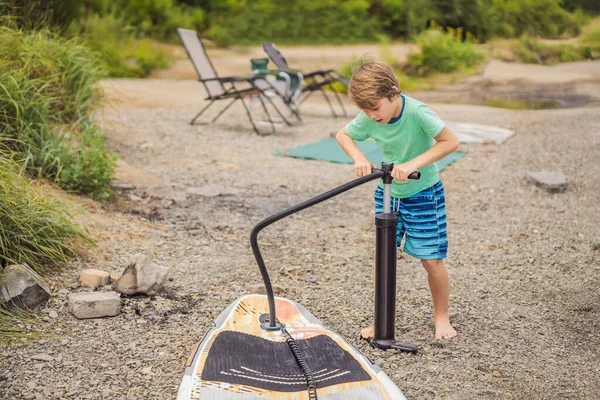 男の子はビーチでポンプでサーフボードを膨らませます。幸せな家族の子供時代の生活。夏の自然屋外個々のウォータースポーツ活動。健康的なライフスタイルのコンセプト — ストック写真