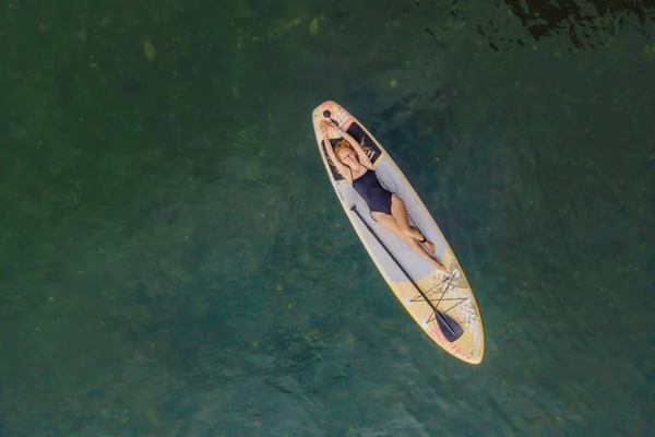 En kvinna ligger på en sup, utsikt från en drönare. Unga kvinnor som har roligt stå upp Paddling i havet. SUP. Rött hår flicka Träning på Paddle Board nära klipporna — Stockfoto