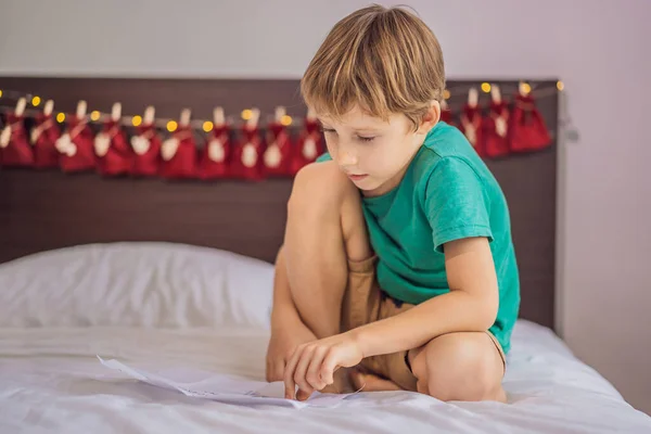 Yatağın üzerinde oturan gülümseyen çocuk takvimden ilk hediyeyi alıyor. Kış mevsimi geleneği. Boşluğu kopyala — Stok fotoğraf
