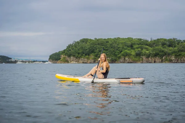 Junge Frauen haben Spaß beim Stand Up Paddling im Meer. SUP. Mädchen mit roten Haaren trainiert auf Paddelbrett in der Nähe der Felsen — Stockfoto